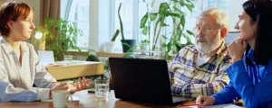 Attorney in white blouse consulting with a senior man in a plaid shirt and a woman in blue at a table with a laptop, representing a car wreck attorney-client meeting.