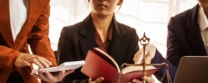 Three attorneys in business attire are consulting over a red law book with justice scales and documents discussing a financial settlement for a personal injury case.