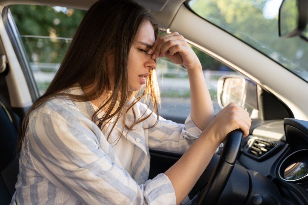 A young woman with long brown hair in a striped shirt, sitting in the driver's seat, looking distressed and holding her forehead after a car accident.