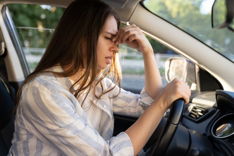 A young woman with long brown hair in a striped shirt, sitting in the driver's seat, looking distressed and holding her forehead after a car accident.