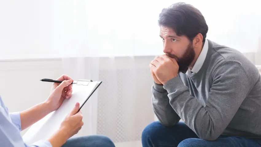 A distressed man is sitting hunched forward with his hands clasped near his face while a clinician takes notes representing someone seeking treatment for PTSD following a car accident in Atlanta.