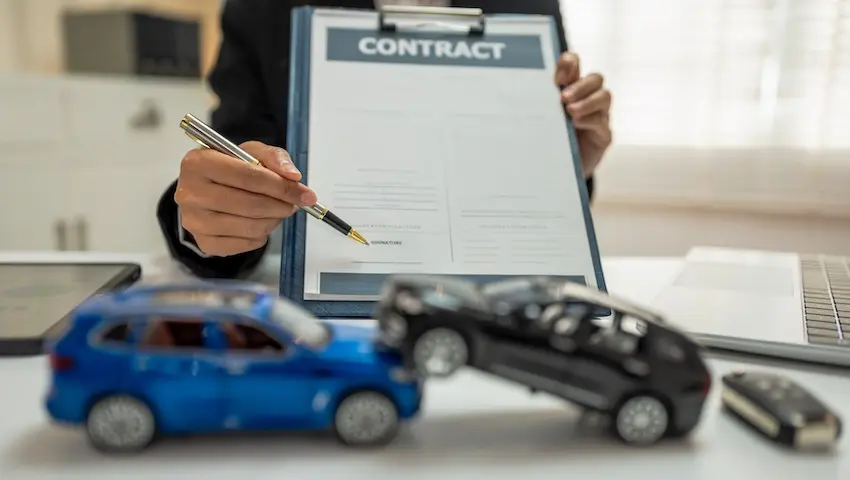 A woman in a black suit holds up a clipboard with a contract document and points to the signature line with a gold-tipped pen while two toy cars staged in a collision sit on the desk in the foreground illustrating the pressure accident victims may feel to sign a first settlement offer before consulting an attorney at Amircani Law.
