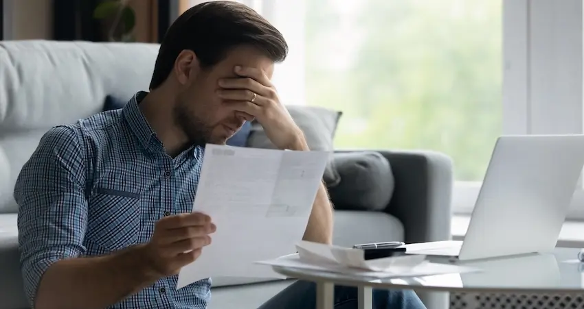 A distressed man holds his head in his hand while reading a document at his home desk beside a laptop- reacting to a denial letter received after filing a claim against an at-fault driver's insurance company following an accident in Metro Atlanta.